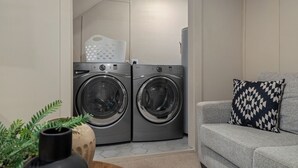 A laundry room with two front-loading washing machines, a grey sofa with a patterned pillow, and a potted plant. - Gothic Garden Bright, Cozy Escape with Modern Touches in Historic Bryan (Bryan)