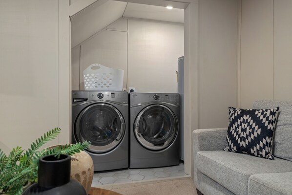 Une buanderie équipée de deux machines à laver à chargement frontal, d’un canapé gris agrémenté d’un coussin à motifs et d’une plante en pot.