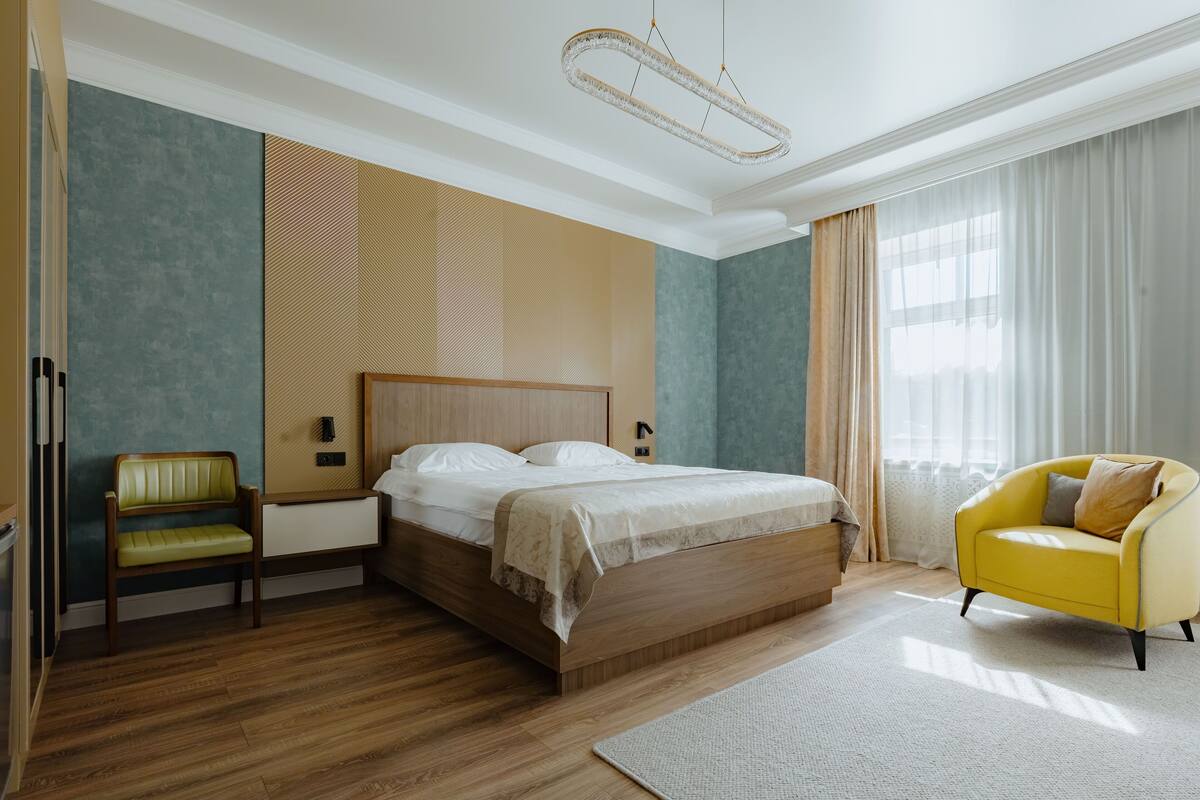 A modern bedroom with a wooden bed, a green upholstered bench, a yellow armchair, and a large window with sheer curtains.
