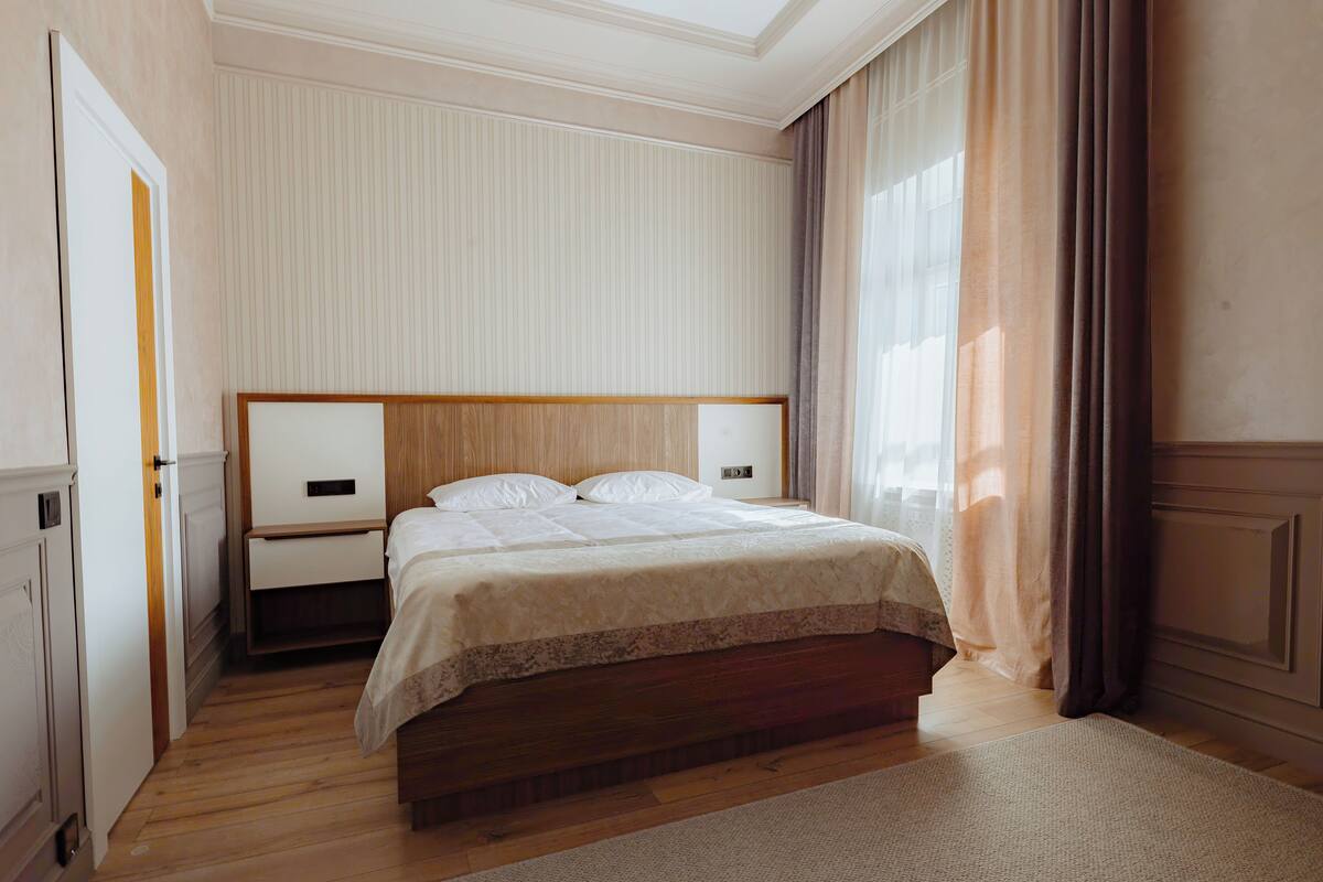 A bedroom with a wooden bed, white bedding, a nightstand, a bedside table, and a large window with curtains.
