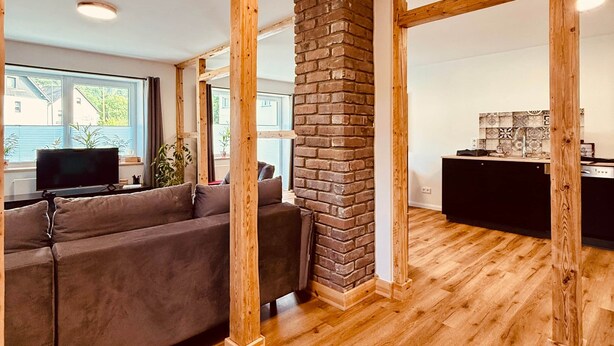 A spacious living area with a wooden floor, a brick accent wall, and a modern kitchen in the background. - Fabian's vacation home / Rurseliebe (Simmerath)