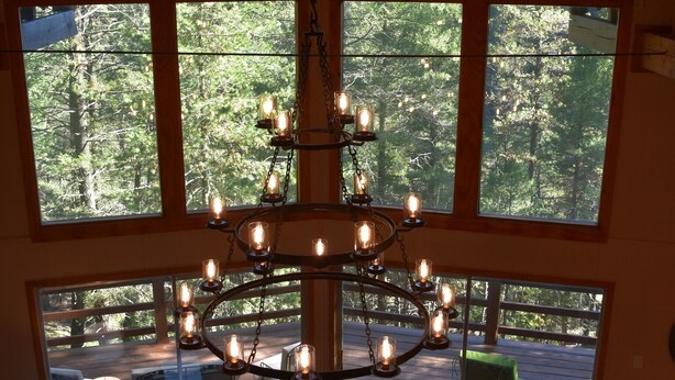 A spacious room with a large chandelier, wooden flooring, and a view of a deck and forest through large windows. - Cloudcroft Hideaway - No Neighbors, Just Nature! Modern Cabin Vibe and SAUNA! (Cloudcroft)