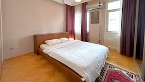 Une chambre avec un lit en bois, une tête de lit blanche, un parquet à motifs en chevrons, un luminaire suspendu et une fenêtre avec des rideaux.