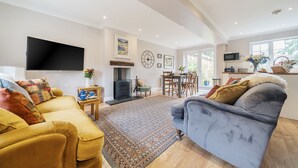 Een woonkamer met een bank, een grijze fauteuil, een flatscreen televisie, een open haard en een eethoek met een tafel en stoelen.