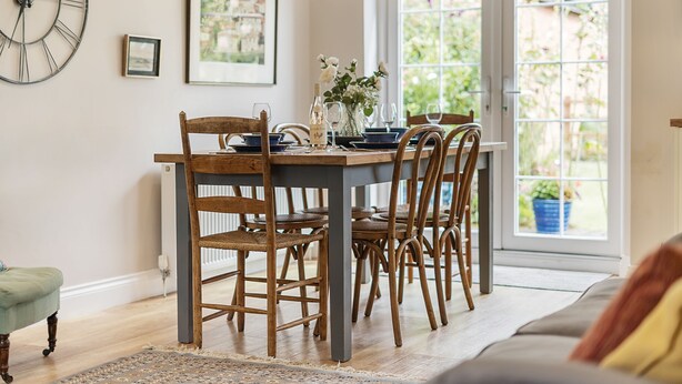 Een eetgedeelte met een houten tafel en stoelen, een grote wandklok, lijstjes met foto's en een raam met uitzicht op groen.