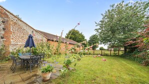 Een bakstenen huis met een stenen terras, een houten schutting en een tuin met bloemen en een tafel met stoelen.