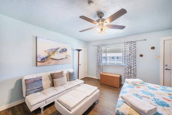 A bedroom with a bed, a sofa, a ceiling fan, a window with patterned curtains, and a painting on the wall.