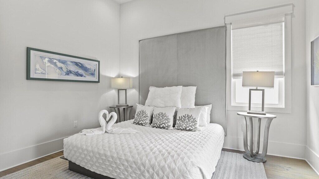 A modern bedroom with a large bed, a grey headboard, a framed artwork on the wall, and a window with a view.