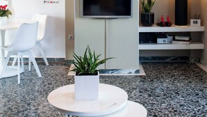 Un intérieur moderne avec un téléviseur à écran plat fixé au mur, une table ronde agrémentée d’une plante en pot, des chaises blanches et un élément décoratif mural.