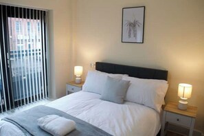 Ein ordentlich bezogenes Bett mit weißen Leinen, zwei Nachttische mit Lampen, ein gerahmtes Bild an der Wand und Blick ins Freie durch ein Fenster mit Jalousien.