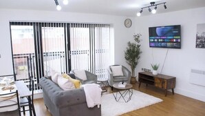 Un soggiorno moderno con un divano grigio, un piccolo tavolino rotondo e un tappeto bianco. C'è una TV montata a parete e una pianta su un tavolino in legno.
