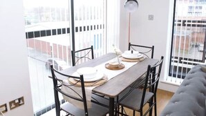 Zona pranzo con tavolo apparecchiato per due, lampada da terra e vista sulla città attraverso ampie finestre.
