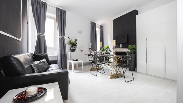 A modern living room with a black leather sofa, a dining area with a white table and chairs, and a kitchen area with a TV and cabinets. - Babuino Luxury Design Apartment (Roma)
