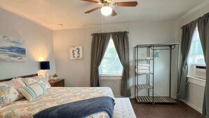 Ein Schlafzimmer mit Bett, einer Sitzbank, Deckenventilator, Fenster mit Vorhängen, Wandregal und einem Gemälde.