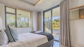 A modern bedroom with a large bed, a sliding glass door to a balcony, and a view of greenery outside.