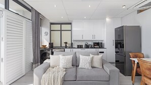 Een moderne keuken met witte kastjes, een grijze bank met decoratieve kussens en een houten eettafel.