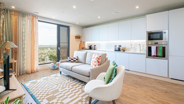 A modern living room with a sofa, armchair, and a view of the city through large windows. - The Eades Laura Perryman 1 Bed (London)