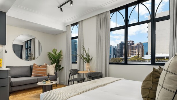 Una habitación de hotel moderna con una cama grande, un sofá, una mesita redonda y vistas a la ciudad a través de amplios ventanales.