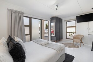 A modern hotel room with a large bed, a flat-screen TV, a wardrobe, and a view of the outside. - San Norina Suite | Blouberg (Cape Town)