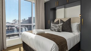 Ein modernes Hotelzimmer mit einem großen Bett, einem Kleiderschrank und Blick auf die Stadt durch große Fenster.
