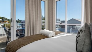 Ein Hotelzimmer mit einem großen Bett, einem Balkon mit Tisch und Stühlen sowie Blick auf die Stadt durch die Fenster.