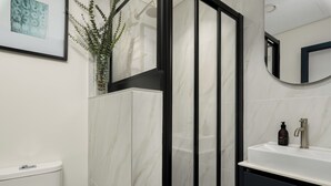 A modern bathroom with a glass-enclosed shower, a white sink with a dark cabinet, and a framed artwork on the wall. - Modern Studio in The Valour | De Waterkant Luxury (Cape Town)