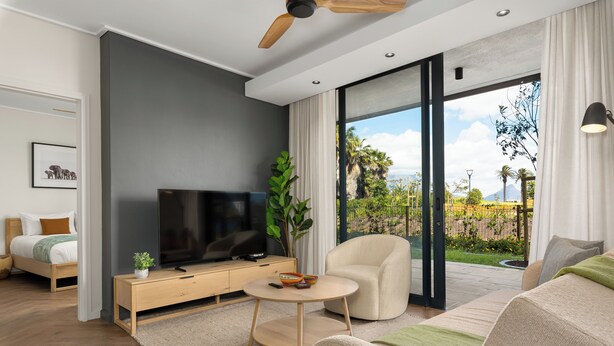 A modern living room with a flat-screen TV, a wooden coffee table, a beige sofa, and a large sliding glass door leading to a balcony with a view of greenery and mountains.