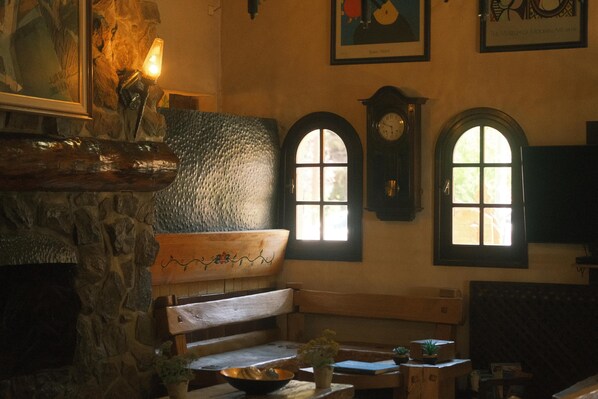 A cozy interior with a stone wall, a wooden bench, and a fireplace. There are two arched windows with a clock above them. - La Posta del Cazador (San Martín de los Andes)