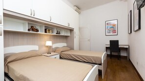 Een slaapkamer met twee bedden, een bureau en een stoel. Er is een plank met decoratieve objecten en een ingelijst beeld aan de muur.