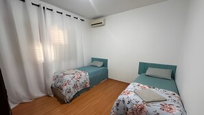 Ein Zimmer mit zwei Einzelbetten, einem Fenster mit weißen Vorhängen und einer Klimaanlage an der Wand.