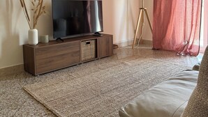 A modern living room with a flat-screen TV on a wooden console, a floor lamp, and a large window with sheer curtains.