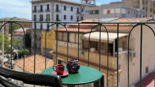 A balcony with a green table, a red cup, and a colorful glass ornament. In the background, there are buildings with a mix of architectural styles.