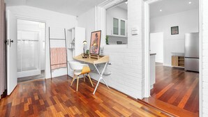 A modern interior with a wooden floor, white brick walls, a desk with a chair, and a kitchen area visible through an open door. - The Ultimate Bedford Modern Stay (Bedford)