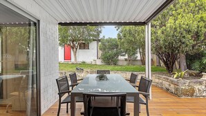 An outdoor patio with a rectangular table and chairs, a wooden deck, and a view of a garden and a building with a red door. - The Ultimate Bedford Modern Stay (Bedford)