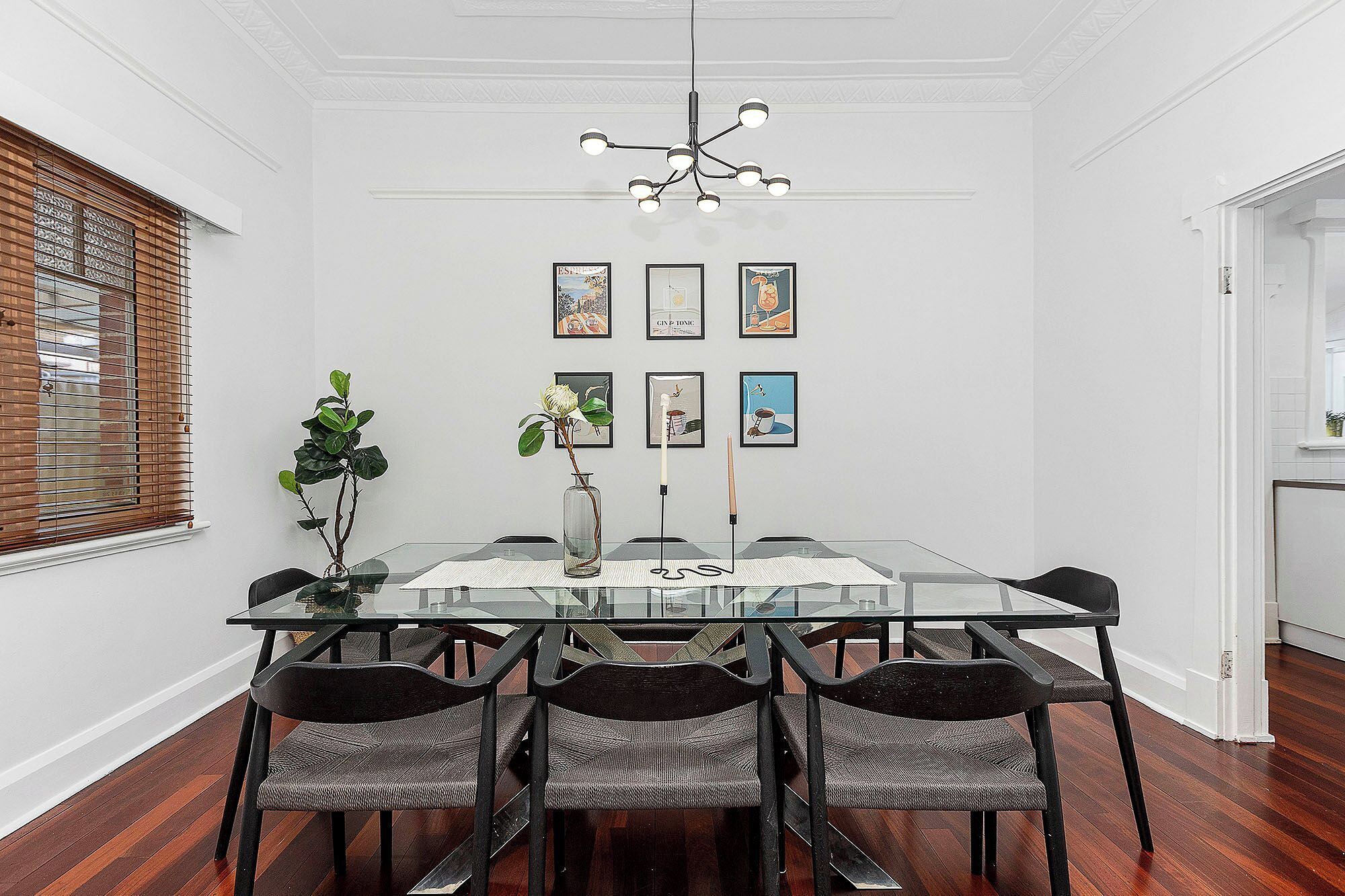 Gather around this elegant glass-top dining table, featuring a stylish gallery wall of art, a modern chandelier, and rich wooden floors.