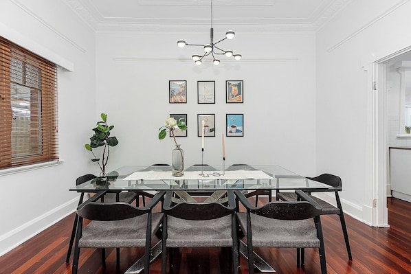 A dining area with a glass table, six chairs, a vase with flowers, and framed artwork on the wall.
