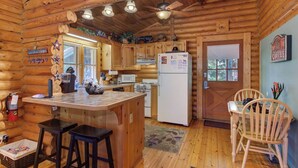 A log cabin interior with a kitchen area, wooden bar stools, a refrigerator, and a dining table with chairs.