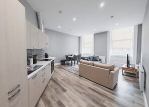 A modern kitchen with white cabinets, a dining area with a table and chairs, and a beige sofa in a living area with large windows. - Edinburgh Castle Suite (Edinburgh)