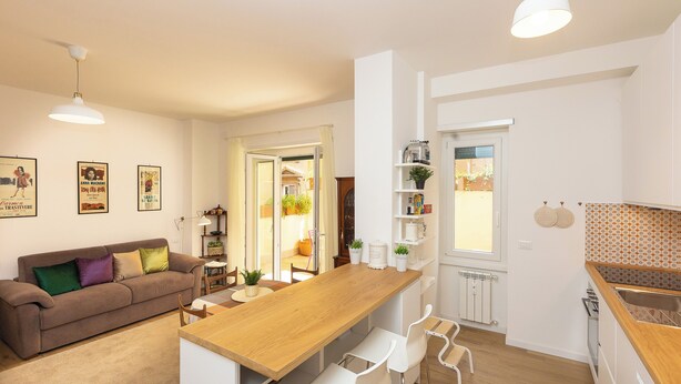A modern kitchen with a wooden table, white chairs, and a built-in oven. There is a white sofa with colorful cushions in the living area, and a dining table with chairs. The kitchen has white cabinets and a window with a view of the outside. - Rental in Rome Annietta Apartment (Rome)