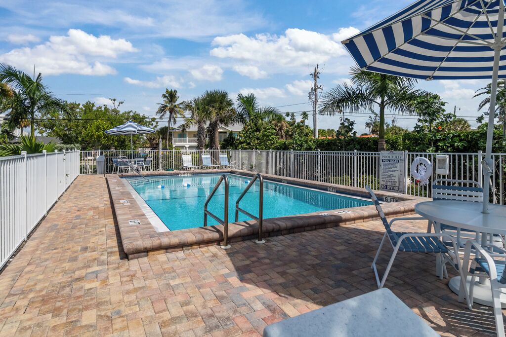 A swimming pool with a white fence, poolside chairs, and a table.