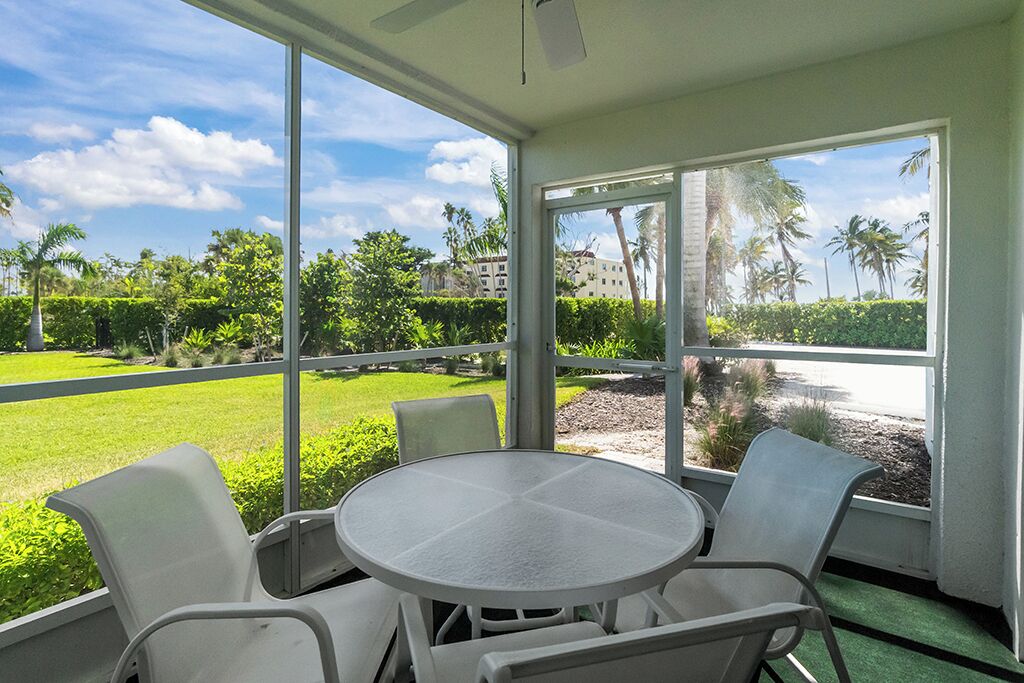 A covered outdoor seating area with a round table and four chairs, overlooking a well-maintained lawn and palm trees.