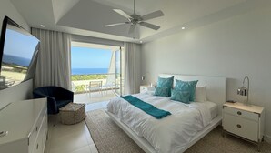 Een moderne slaapkamer met een groot bed, een plafondventilator, een televisie aan de linkerkant en een balkon met uitzicht op de oceaan.