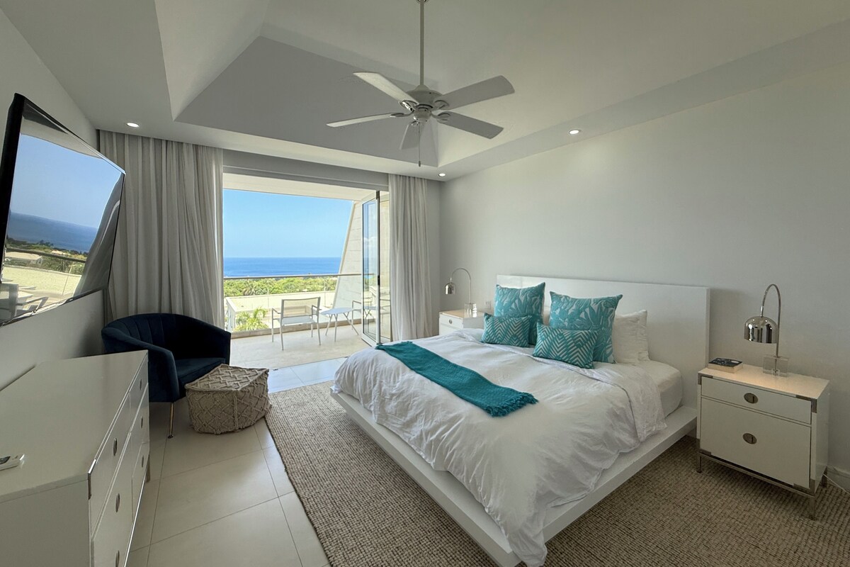 Une chambre moderne avec un grand lit, un ventilateur de plafond, un téléviseur sur le côté gauche et un balcon offrant une vue sur l’océan.