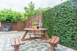 A wooden picnic table with benches, a bottle of wine, and a glass of water on a patio with a checkered tile floor.