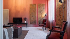 Un salon comprenant un téléviseur, un fauteuil rouge, une chaise noire, une petite table avec un vase et deux grandes peintures murales.