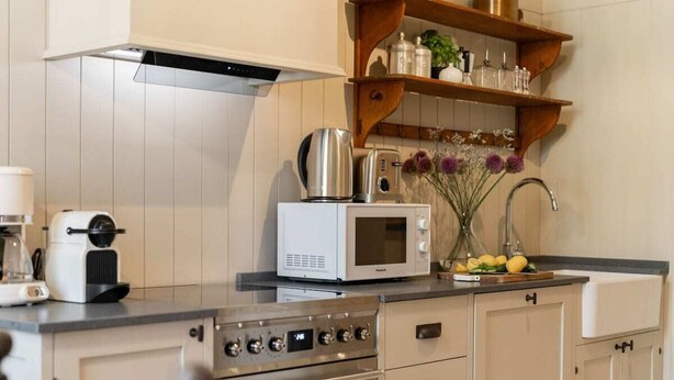 Une cuisine équipée d’un four à micro-ondes, d’un grille-pain et d’une cuisinière. Des étagères en bois, situées au-dessus du plan de travail, servent à ranger des bocaux et un vase avec des fleurs.