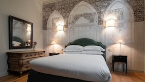 Une chambre à coucher avec un grand lit, une commode et un miroir. On y trouve des appliques murales et un papier peint à motifs.