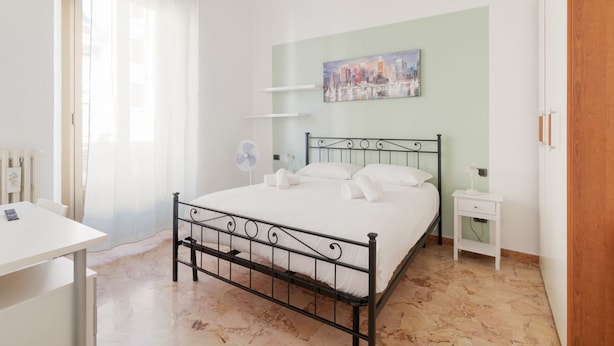 A bedroom with a bed, a bedside table, a chair, a desk, a fan, and a painting on the wall.