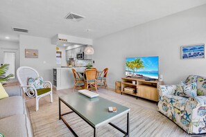 A modern living room with a dining area, kitchenette, and a TV displaying a beach scene.