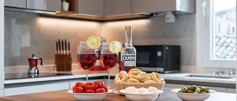 A kitchen counter with a bottle of Campare, two glasses of red wine, a bowl of cherry tomatoes, a basket of bread, and a bowl of olives.